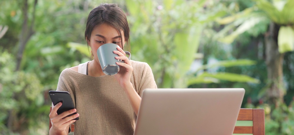 Woman sat at a laptop drinking a hot drink and reading phone